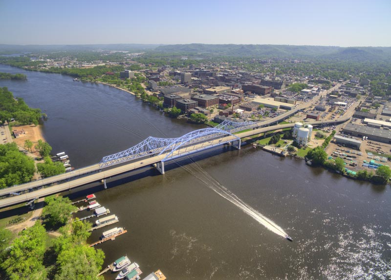 Wide view of La Crosse’s blue bridge across the Mississippi. A single motorboat glides down the river with a long white stream of wake behind it.