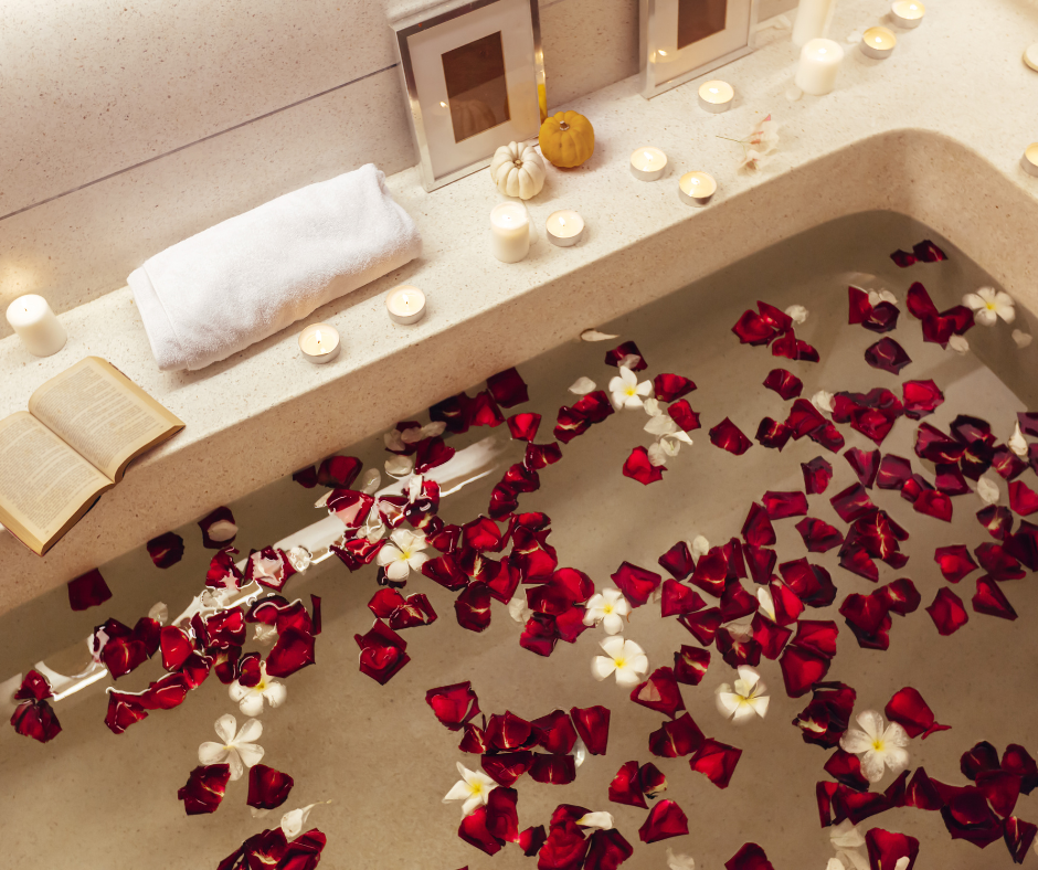 a cream colored bathtubs filled with roses and other flowers surrounded by candles, a book, and a towel