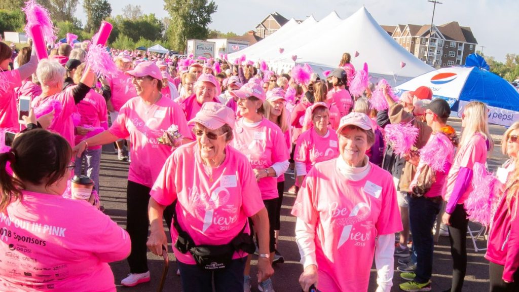 Participants in bright pink shirts walk together during the Steppin’ Out in Pink event, surrounded by cheering crowds and vendor tents