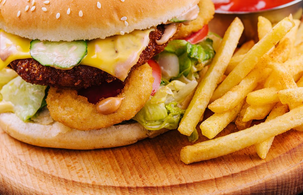Close-up of a cheeseburger topped with lettuce, tomato, cucumber slices, melted cheese, onion rings, and special sauce, served with a side of seasoned French fries on a wooden plate