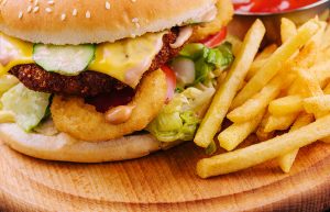 Close-up of a cheeseburger topped with lettuce, tomato, cucumber slices, melted cheese, onion rings, and special sauce, served with a side of seasoned French fries on a wooden plate