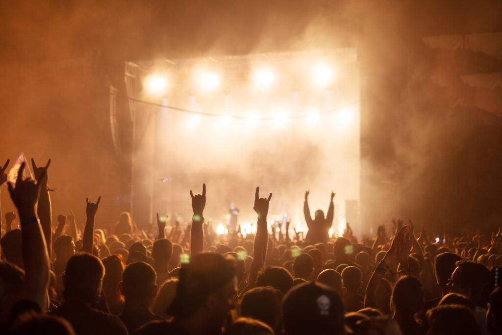 A vibrant crowd at a nighttime outdoor rock concert, with silhouetted fans raising their hands in the air as bright stage lights and fog illuminate the scene in front of a large performance stage.