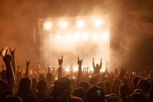 A vibrant crowd at a nighttime outdoor rock concert, with silhouetted fans raising their hands in the air as bright stage lights and fog illuminate the scene in front of a large performance stage.