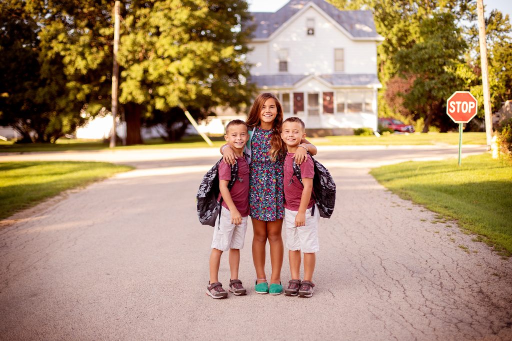 Back-to-school photo of three happy children in a neighborhood street, wearing backpacks and smiles. Promotes La Crosse’s Back2School Program providing free school supplies.