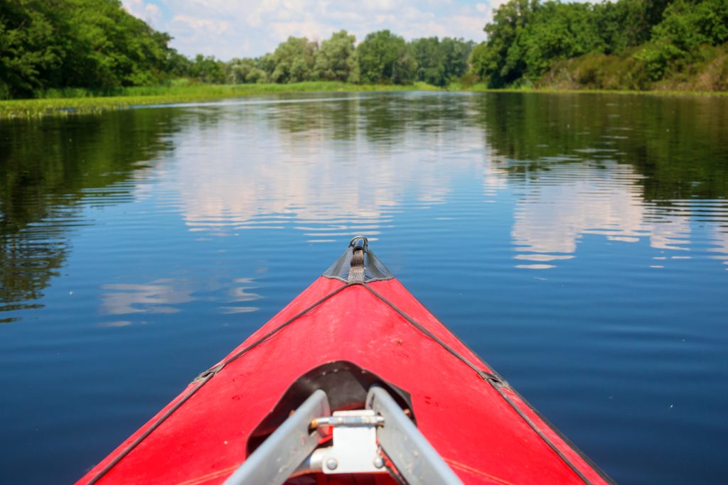 View from a red kayak on calm water with green trees and blue sky reflections, La Crosse, Wisconsin