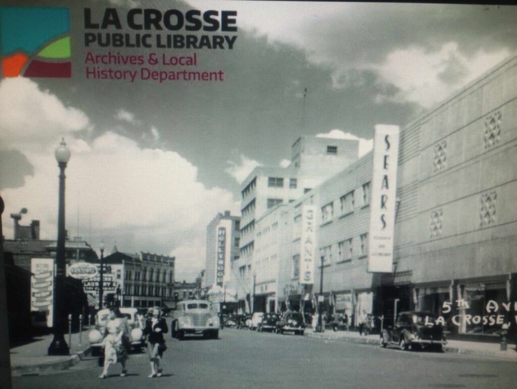 Black-and-white historic photo of 5th Avenue in downtown La Crosse, Wisconsin, showing Sears and other storefronts with vintage cars and pedestrians.