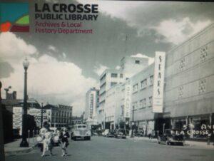 Black-and-white historic photo of 5th Avenue in downtown La Crosse, Wisconsin, showing Sears and other storefronts with vintage cars and pedestrians.