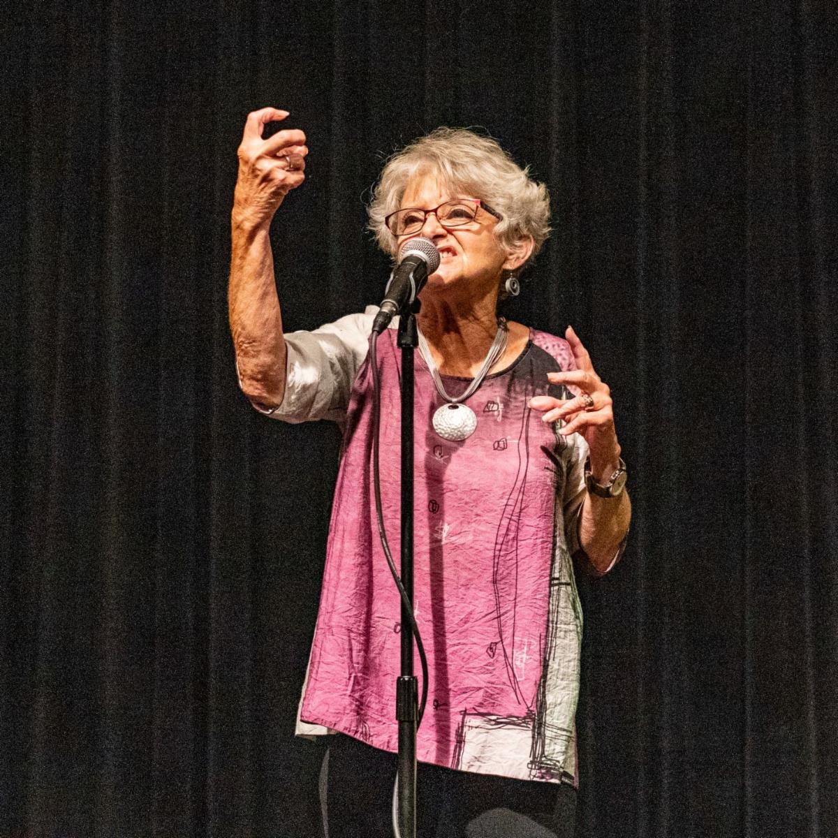 A Bluff Country Tale Spinners storyteller performs on stage at the Pump House in La Crosse, Wisconsin, raising her hand expressively while speaking into a microphone.