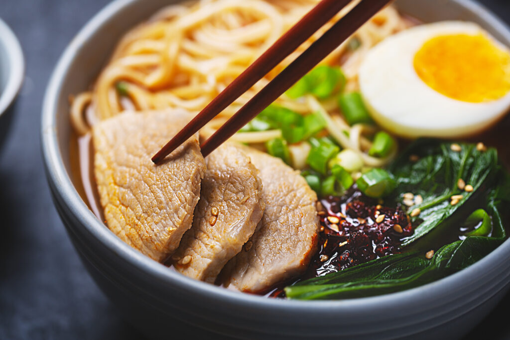 Close-up of a bowl of Japanese ramen with sliced pork, noodles, green onions, leafy greens, chili oil, and a soft-boiled egg held with chopsticks.