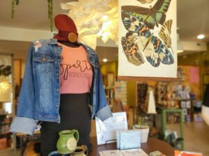 Display inside KEPS Sparta featuring a mannequin wearing a &ldquo;Sparta Wisconsin&rdquo; shirt, denim jacket and hat, surrounded by gift items, a green mug, and a butterfly wall art print in the background.