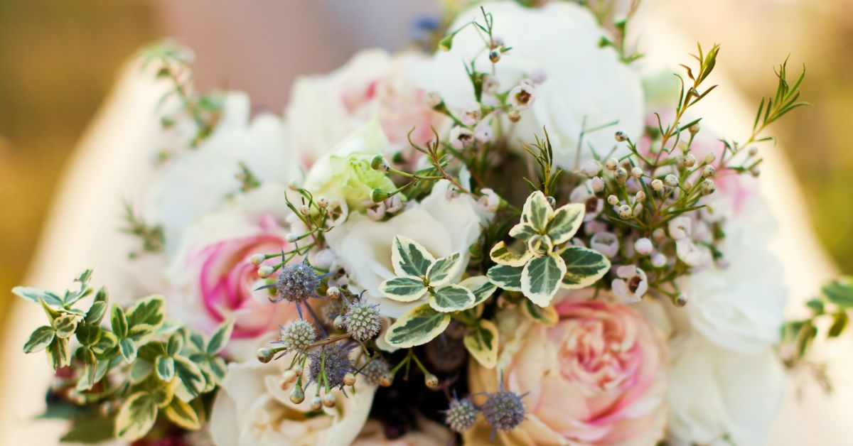 Close-up of a soft, romantic bridal bouquet featuring white and blush pink roses, small white filler flowers, and green variegated foliage, arranged in a natural, airy style.